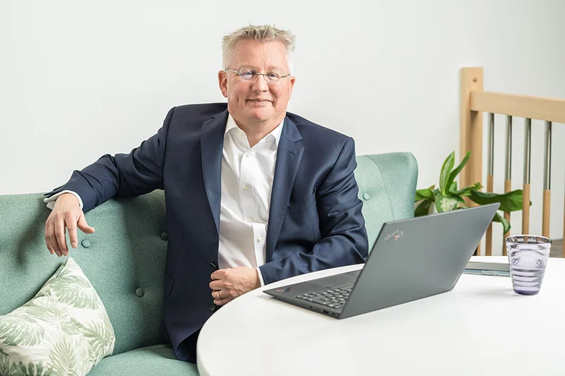 Fritz Naumann in einem blauen Anzug auf einem Sofa sitzen, vor sich einen offenen Laptop